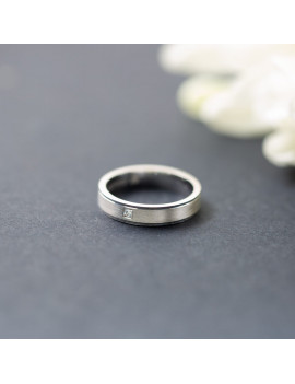 Anillo de oro blanco con diamante princesa y acabado satinado sobre un fondo oscuro junto a una flor blanca.
