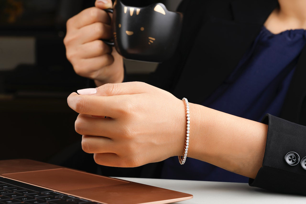 brazo de mujer elegante con pulsera tenis de diamantes. Sostiene una taza de café mientras mira el ordenador y en la otra mano brillan las gemas del brazalete riviere de diamantes